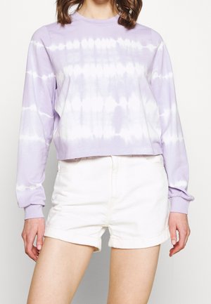 Kobieta w lawendowo-białej koszulce z długim rękawem ze wzorem tie-dye, zestawiona z białymi, wysokim stanem i podwiniętymi szortami.