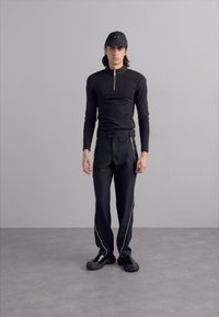 Haut noir à manches longues avec un col à demi-zip, associé à un pantalon noir avec des zips latéraux en argent et des chaussures noires épaisses.