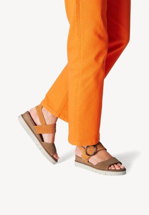 Benen in feloranje broeken die bruine open sandalen met dikke witte zolen en een grote ronde gesp aan de zijkant dragen.