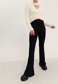 Pull en tricot blanc duveteux avec un élément en forme de cœur à l'avant, associé à un pantalon noir à larges côtes évasé et des chaussures à plateforme noires épaisses.