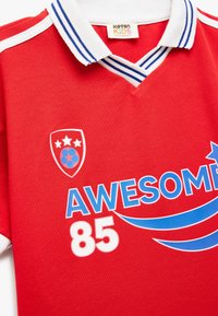 Camiseta de fútbol infantil roja con cuello blanco y rayas azules. Presenta un logo impreso, la palabra "ASOMBROSO" y el número "85". Tejido texturizado.