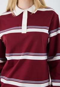 Polo à manches longues bordeaux avec un col blanc, featuring des rayures horizontales blanches et bleues sur la poitrine. Détail de patte de boutonnage à quatre boutons.