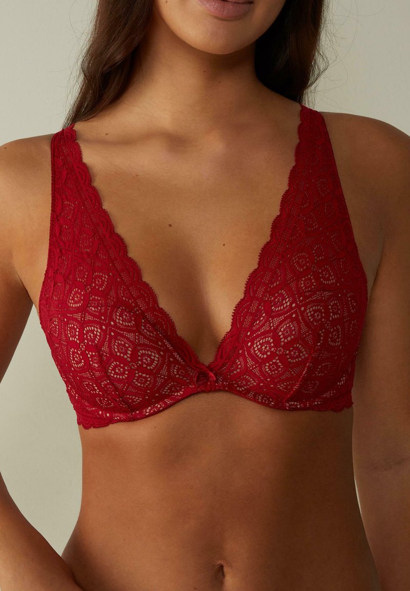 Červená krajková bralette s hlubokým výstřihem do V, se scalloped okraji a centrálním mašlí. Design obsahuje květinový vzor s průsvitnými částmi.