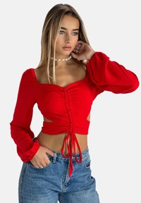 Top rosso corto con maniche lunghe a sbuffo, dettaglio arricciato nella parte frontale e ritagli laterali. Abbinato a jeans in denim blu a vita alta.