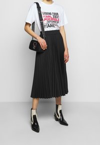 Bílý tričko s černým a červeným textem, černá plisovaná midi sukně, malá černá taška a černé kotníkové boty s bílými akcenty.