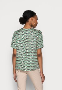 Short-sleeve shirt in light green fabric featuring a geometric triangle pattern in dark green, navy, and beige. Flowy design with a back pleat.