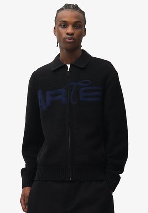 Giovane uomo con capelli intrecciati che indossa un maglione nero con zip e un motivo grafico blu sul petto su sfondo bianco.
