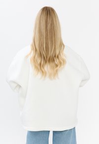 Oversized biały sweter z opadającymi ramionami, miękką teksturą i ribbanym wykończeniem. Połączony z jasnoniebieskimi dżinsami.