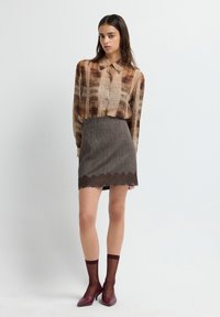 Donna in piedi su uno sfondo bianco, indossa una blusa marrone a fantasia, una gonna grigia con orlo in pizzo, calze trasparenti borgogna e tacchi abbinati.