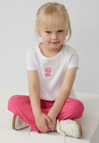 Witte t-shirt met korte mouwen en roze schelpenborduurwerk, gecombineerd met roze ribbroek en witte sneakers met beige zolen.