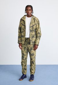 �Φόρμα καμουφλάζ με τζάκετ και παντελόνι σε ελιά πράσινο χρώμα με σκούρες λεπτομέρειες, διαθέτει φερμουάρ στο μπροστινό μέρος, ρυθμιζόμενα κορδόνια και χαλαρή εφαρμογή.