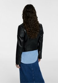 Giubbotto da motociclista in pelle nera con design trapuntato sulle spalle e dettagli con zip. Indossato sopra una camicia di denim azzurro chiaro e una gonna lunga.