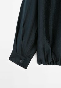 Chemise noire à manches longues avec bouton de manchette et ourlet élastiqué, tissu texturé, présentée sur fond blanc.