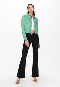 Giacca di jeans corta verde menta con due tasche frontali, abbinata a jeans neri a zampa d'elefante e sandali trasparenti con tacco. Sotto una maglietta crop bianca aderente.