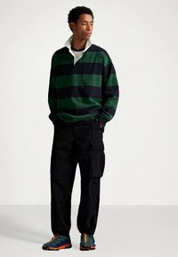 Πολο με μακριά μανίκια σε πράσινες και ναυτικές ρίγες, με λευκό γιακά, συνδυασμένο με μαύρο παντελόνι cargo και πολύχρωμα αθλητικά παπούτσια.