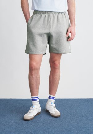 Graue Baumwollshorts mit elastischem Bund, seitlichen Taschen und einem kleinen Logodetail. Kombiniert mit weißen Sneakern und blau gestreiften Socken.