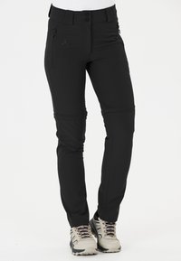 Pantalons d'extérieur noirs en tissu extensible, avec une poche zippée, un système de fermeture à bouton et des poignets ajustables. Associés à des chaussures de randonnée de couleur claire.