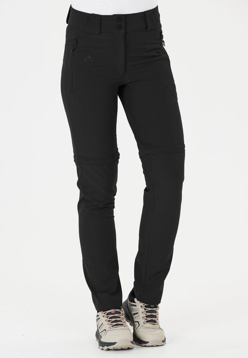 Pantalons d'extérieur noirs en tissu extensible, avec une poche zippée, un système de fermeture à bouton et des poignets ajustables. Associés à des chaussures de randonnée de couleur claire.
