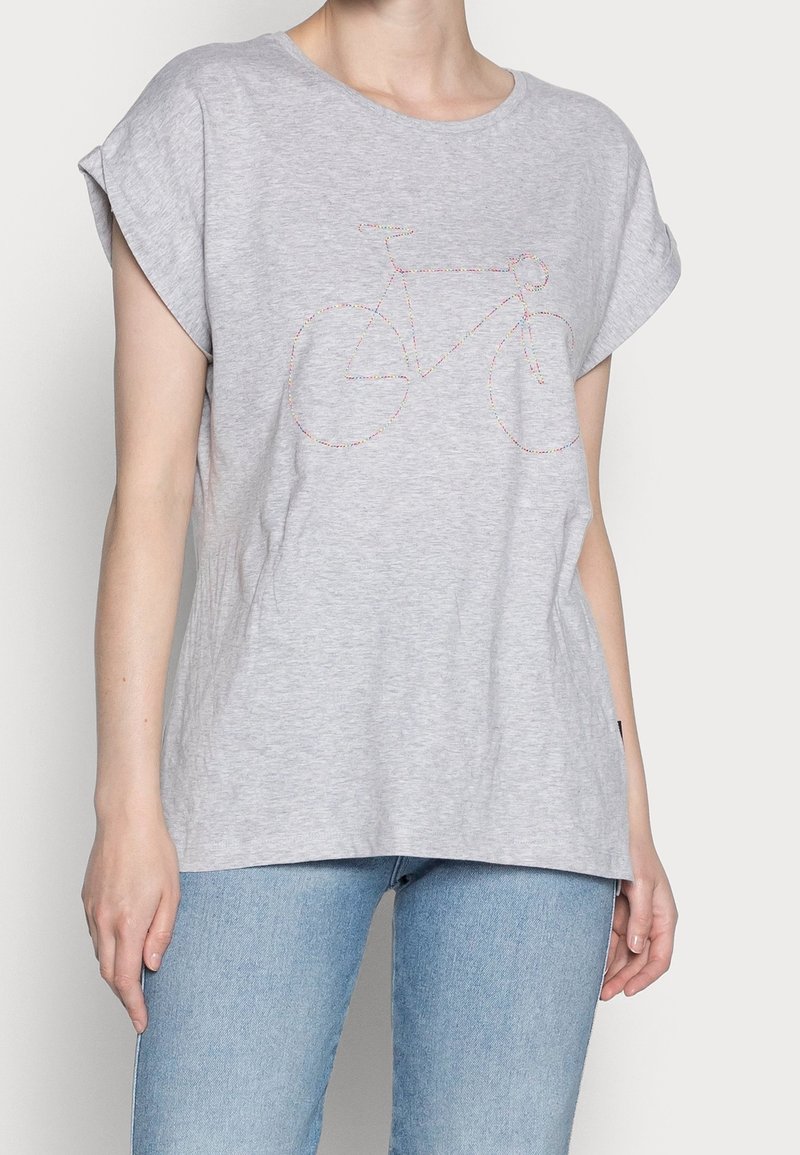 Femme portant un t-shirt gris à manches courtes avec un design de bicyclette coloré brodé sur le devant, associé à un jean bleu clair.