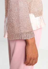 Světle růžový pletený svetr s texturovaným vzorem, volnými rukávy a vrstveným růžovým saténovým podkladem se zlatými akcenty.