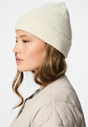 Ribbade, krämfärgade mössa gjord av mjukt material, med en vikbar brätte och en rundad topp, bärs med en ljusfärgad vadderad jacka.