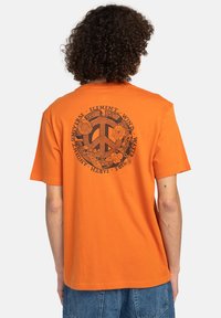 Camiseta de algodón naranja con un símbolo de paz negro decorado con diseños florales y texto alrededor, de manga corta y corte relajado.