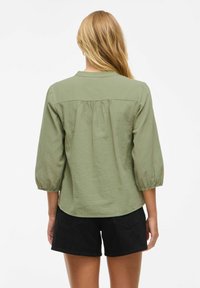 Blusa leggera verde oliva, con maniche 3/4, scollatura rotonda e dettaglio arricciato sul retro, abbinata a pantaloni corti neri.