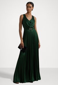 Grön plisserad klänning med djup V-ringning och justerad midja, prydd med ett dekorativt bälte. En svart clutchtvål hålls i ena handen. Slät textur.