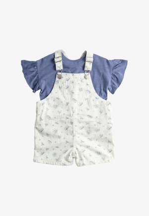 Chemise bleue à manches courtes avec volants, associée à une salopette courte blanche à imprimé floral, avec poche avant et bretelles ajustables.