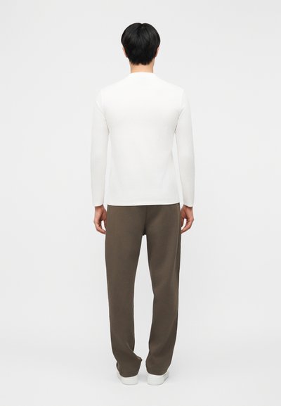 Άνδρας με μαύρα μαλλιά στέκεται με την πλάτη γυρισμένη, φορώντας λευκό μακρυμάνικο πουκάμισο, καφέ παντελόνι και λευκά αθλητικά παπούτσια σε απλό λευκό φόντο.