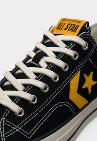 Καμβάς αθλητικού παπουτσιού με μαύρο επάνω μέρος, κίτρινο αστέρι και λογότυπο, λευκά κορδόνια και αντίθετη λευκή ραφή. Κλασική λαστιχένια σόλα.