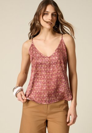 Donna con top smanicato rosso con motivi e pantaloni beige, che tiene l'orlo con la mano destra, sfondo beige.