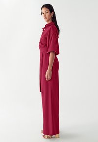 Punainen napitettava mekko, jossa on lyhyet puffihihat, istuva vyötärö ja leveät housunlahkeet. Silkkinen kangas, jossa on vain vähän koristeita. Pitkät, paksupohjaiset sandaalit.