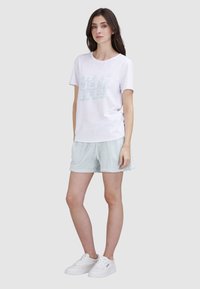 T-shirt blanche avec un graphique "TRUE JEAN BABY" en bleu clair, associée à un short bleu clair. Le mannequin porte des baskets blanches, se tenant sur un fond gris.