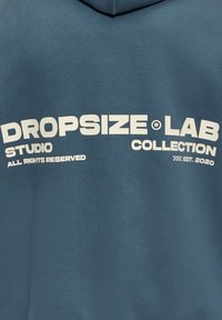 Tissu bleu avec texte imprimé en blanc : "DROPSIZE LAB STUDIO COLLECTION TOUS DROITS RÉSERVÉS ÉTABLI EN 2020."