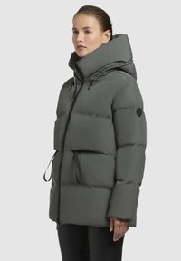 Dikke olijfgroene jas met hoge kraag, verstelbare capuchon en zwarte rits. Heeft trekkoorden en een logopatch op de linkermouw.