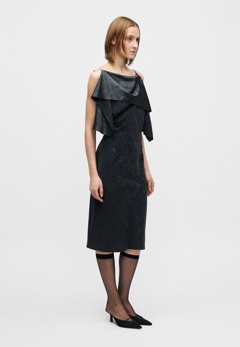 Abito nero con corpetto in raso a strati, tessuto strutturato, design dritto e lunghezza sopra il ginocchio; abbinato a calze nere trasparenti e tacchi a punta.