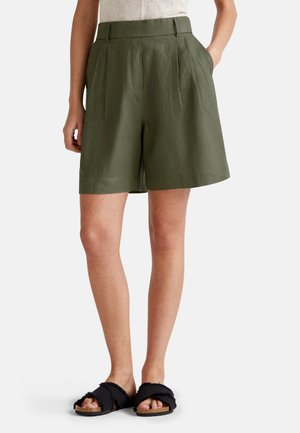 Zielone lniane szorty z wysokim stanem, plisami i bocznymi kieszeniami. Noszone z czarnymi, teksturowanymi klapkami na płaskim obcasie.