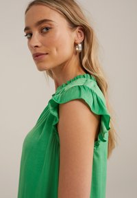Blusa verde con maniche a sbuffo, tessuto morbido e taglio rilassato. Il modello indossa orecchini a cerchio argentati, mostrando un profilo laterale.
