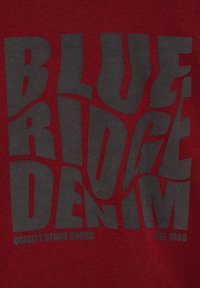 Tessuto rosso con testo bold in nero che legge "BLUE RIDGE DENIM" con un design ondulato; un testo più piccolo dice "QUALITY DENIM GOODS EST. 1986."