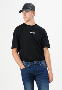 Camiseta de algodón negra con texto japonés, combinada con jeans ajustados de mezclilla azul. El modelo lleva una gorra de cuero negra. Diseño sencillo, aspecto casual.