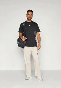 Camisola atlética preta com riscas brancas, calças em azul claro e ténis cinzentos. Modelo segura uma mala de desporto preta com uma marcação branca.