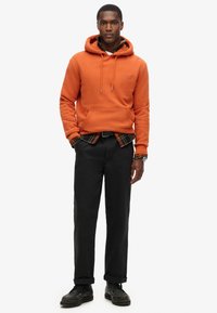Sudadera naranja con capucha y bolsillo frontal, superpuesta sobre una camisa de cuadros, combinada con pantalones negros y zapatos negros con cordones.