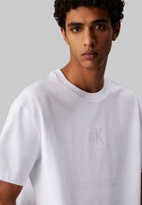 Camiseta de algodón blanca con cuello redondo, que presenta un sutil logo bordado en color a juego en el pecho. Textura suave, corte holgado.