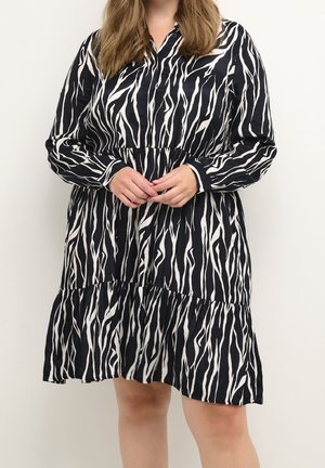 Vrouw draagt een zwart-witte zebraprint lange mouwen jurk met een gelaagde rok, staand tegen een effen lichte achtergrond.