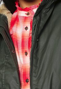 Grön jacka med en krage i päls, lagd över en röd och vit rutiga skjorta med knappar och en volangkrage.