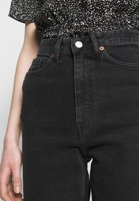 Černé džíny s vysokým pasem, mírně texturované, se pěti kapsami a stříbrným knoflíkovým zapínáním. Tkanina vypadá odolně a trvanlivě.