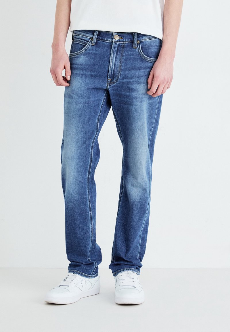 Homme portant un jean bleu délavé et des baskets blanches, debout devant un fond clair uni.