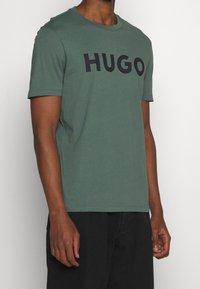Tmavě zelené bavlněné tričko s velkým černým logem "HUGO" na přední straně. Klasický výstřih, krátké rukávy a měkká, hladká textura.