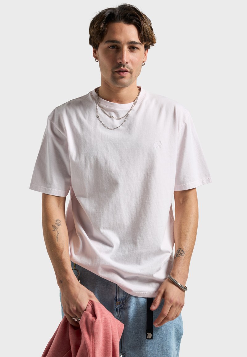 T-shirt en coton rose clair à manches courtes, coupe décontractée, avec un détail de logo subtil. Le mannequin tient un sweat-shirt rouge et porte des chaînes en argent superposées.